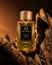 A bottle of Okaasha Jabal Oud extrait de parfum stands on textured wood logs against a warm, dark brown background.