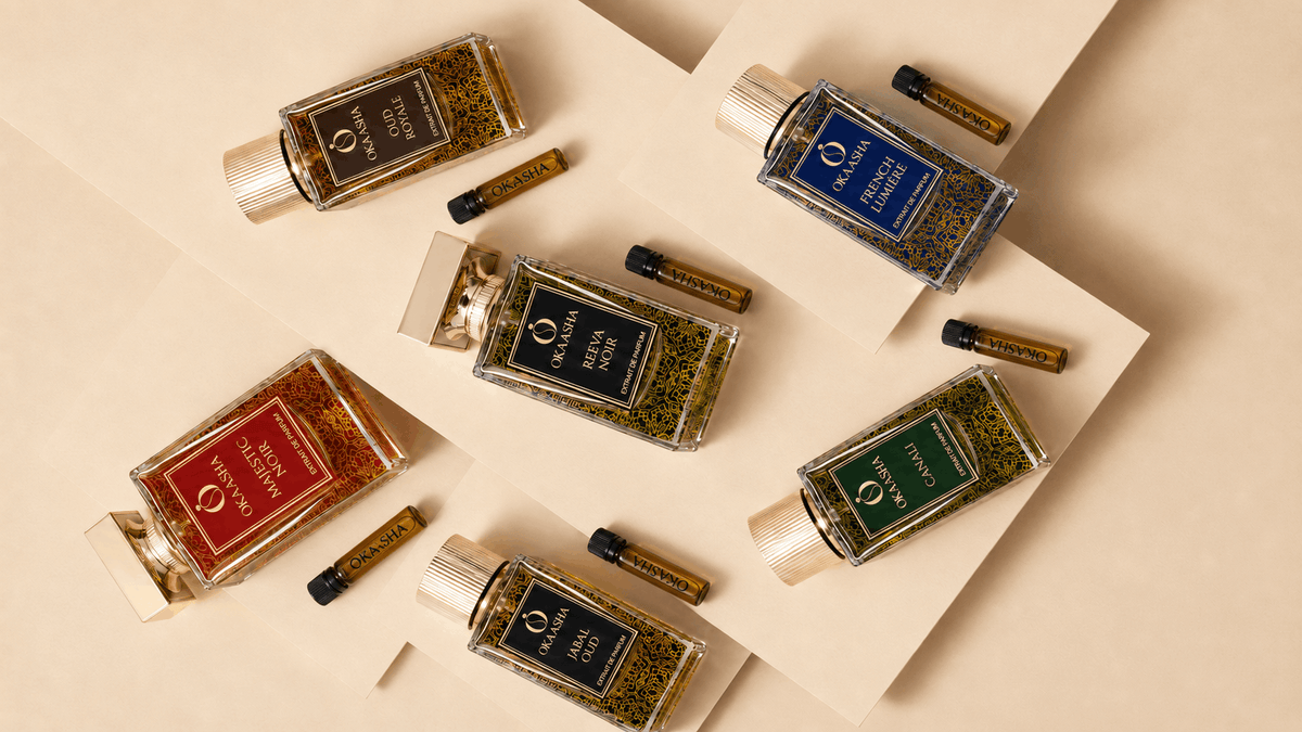 Six glass perfume bottles with gold caps and small matching sample vials arranged on beige geometric paper sheets. The bottles contain amber, green, blue, and red liquids with gold labels, and the brand name 'Okaasha' is visible on the labels.