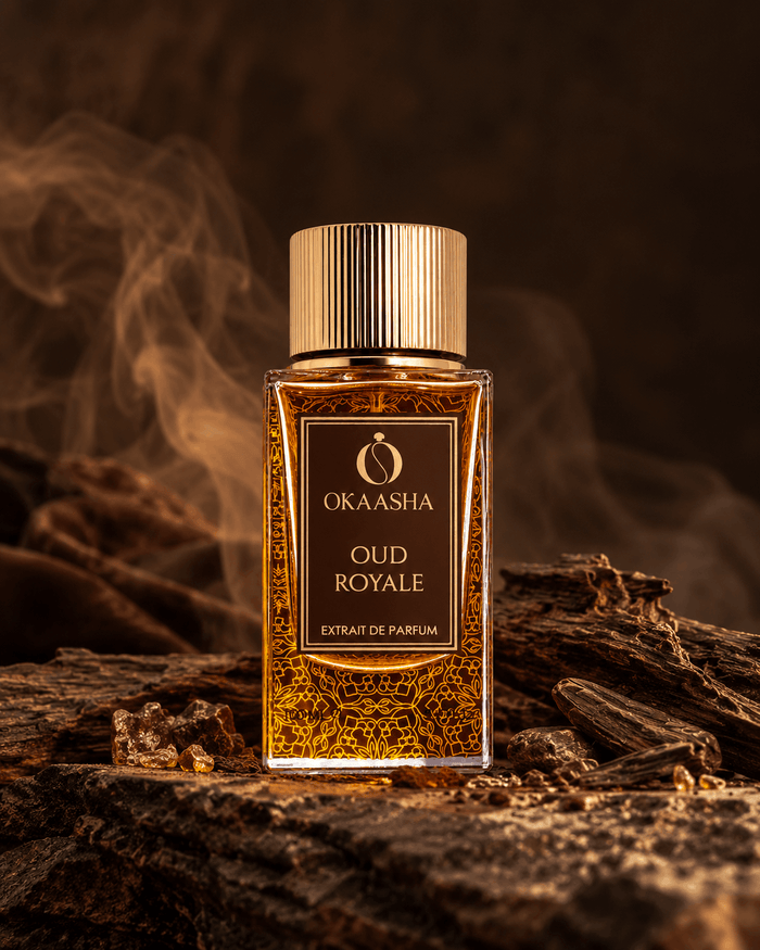 A rectangular glass bottle of Okaasha Oud Royale extrait de parfum with a gold cap, filled with amber-colored liquid and gold patterned flakes, set on a piece of wood with a smoky, dark brown background.