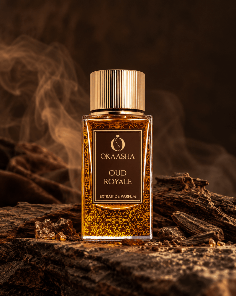 A rectangular glass bottle of Okaasha Oud Royale extrait de parfum with a gold cap, filled with amber-colored liquid and gold patterned flakes, set on a piece of wood with a smoky, dark brown background.