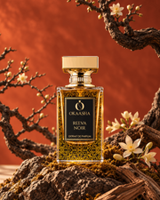 A bottle of Okaasha Reeva Noir extrait de parfum stands on a mossy rock surrounded by branches and small white flowers against a warm orange background.