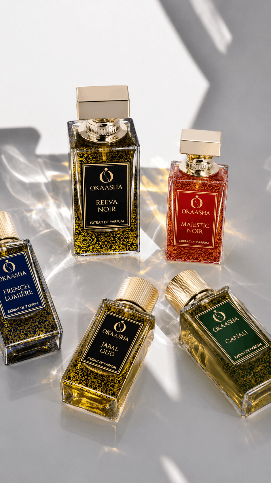 Five Okaasha perfume bottles arranged on a glossy white surface with dramatic diagonal shadows. The bottles are rectangular glass containers with gold caps and colorful labels.